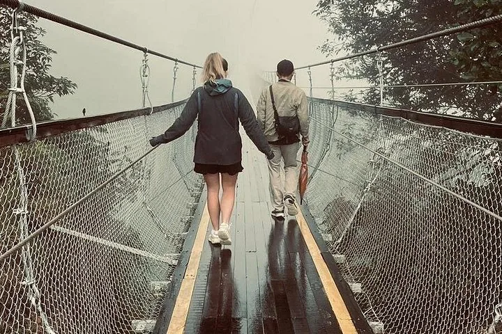 Dos personas alejándose por un puente colgante mojado en un día de niebla.